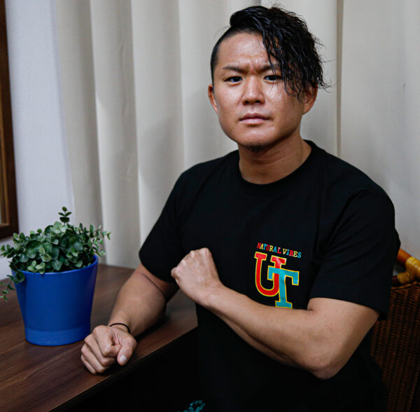 U-T is seated at a table, donning casual clothes, with a potted plant beside him