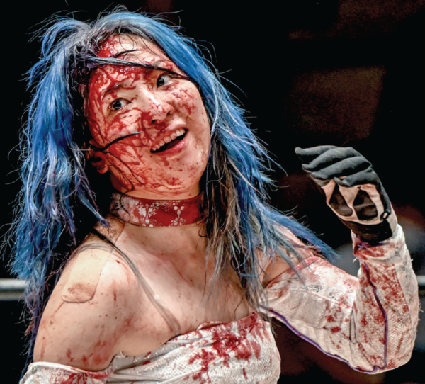 A close-up photo of Risa Sera, who is all smiles despite her face and white ring gear being caked in blood
